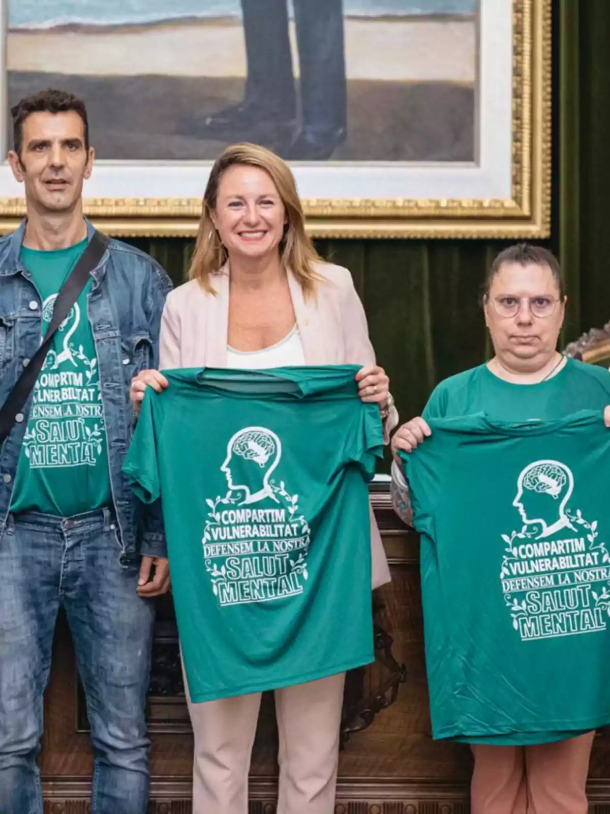 Tres personas posan sonrientes mostrando camisetas verdes con un mensaje a favor de la salud mental en un entorno institucional con un cuadro al fondo. Tres personas posan sonrientes mostrando camisetas verdes con un mensaje a favor de la salud mental en un entorno institucional con un cuadro al fondo.
