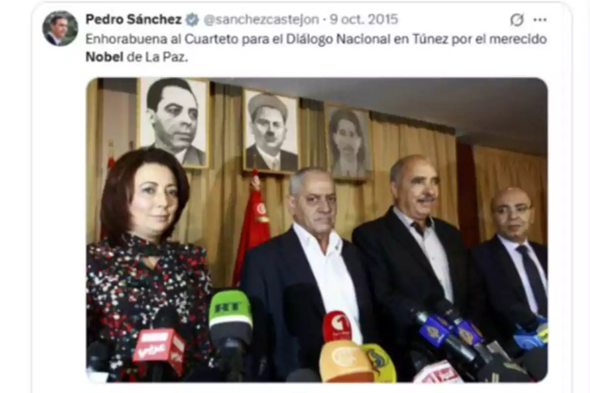 Cuatro personas posan frente a varios micrófonos en una rueda de prensa, con retratos en blanco y negro colgados en la pared detrás de ellos y una bandera roja al fondo.