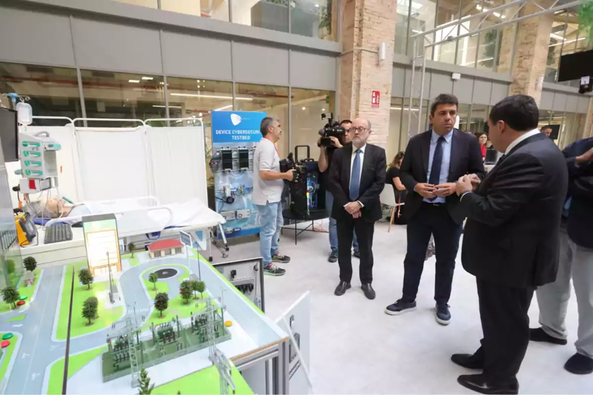 Varios hombres de traje conversan en una sala moderna junto a una maqueta de ciudad y un área de simulación médica, mientras un camarógrafo graba la escena. Varios hombres de traje conversan en una sala moderna junto a una maqueta de ciudad y un área de simulación médica, mientras un camarógrafo graba la escena.