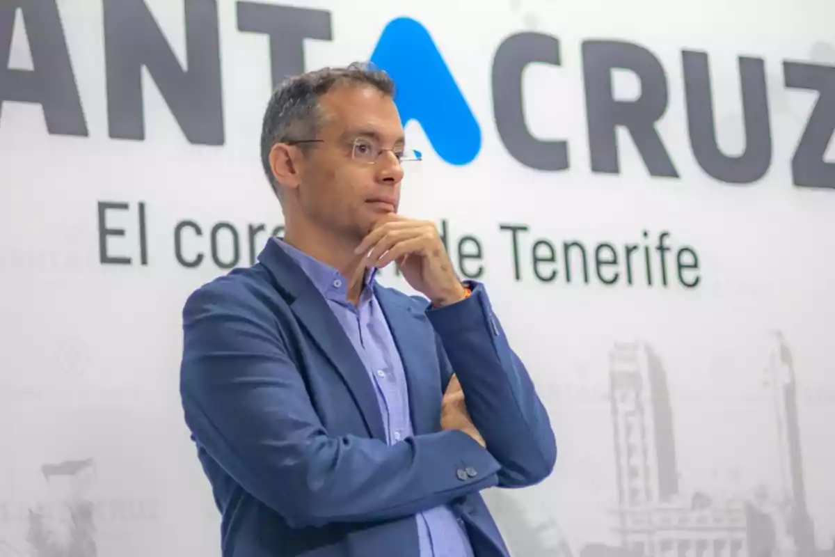 Hombre de pie con gafas y chaqueta azul en actitud pensativa frente a un cartel que dice Santa Cruz El corazón de Tenerife