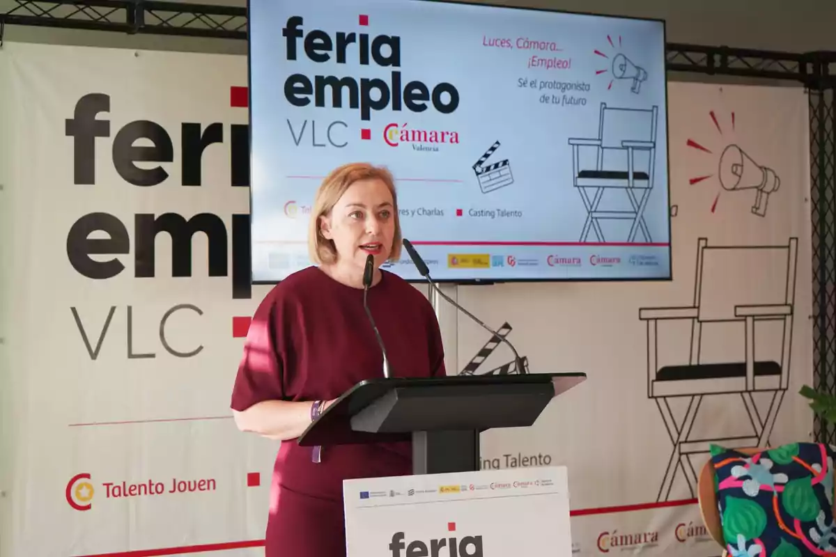Mujer de cabello rubio y vestido burdeos hablando en un podio durante un evento de feria de empleo en Valencia con un fondo que muestra el logo del evento y una pantalla con información sobre la feria Mujer de cabello rubio y vestido burdeos hablando en un podio durante un evento de feria de empleo en Valencia con un fondo que muestra el logo del evento y una pantalla con información sobre la feria