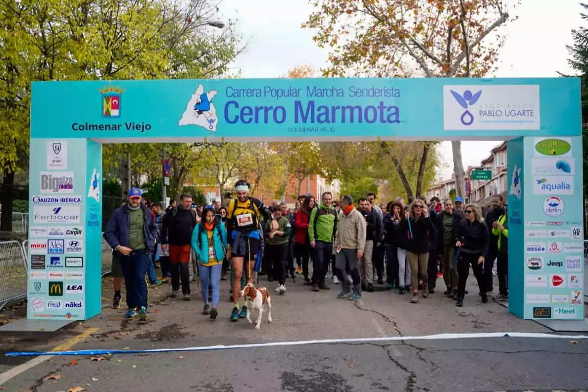 Un grupo de personas y un perro cruzan la línea de salida en una carrera popular de senderismo llamada Cerro Marmota en Colmenar Viejo bajo un arco azul con árboles y casas al fondo