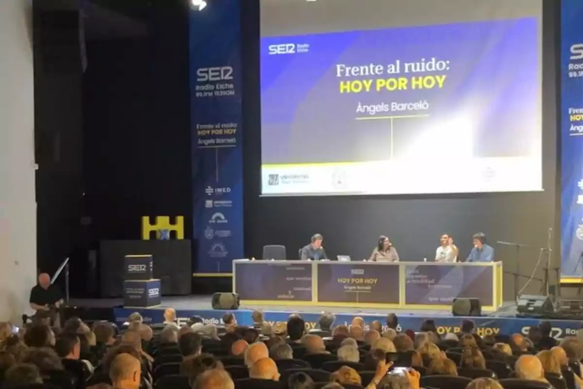 Auditorio lleno de personas asistiendo a una mesa redonda con cuatro ponentes en un escenario, con una pantalla grande al fondo que muestra el título del evento Frente al ruido HOY POR HOY y el nombre de Àngels Barceló