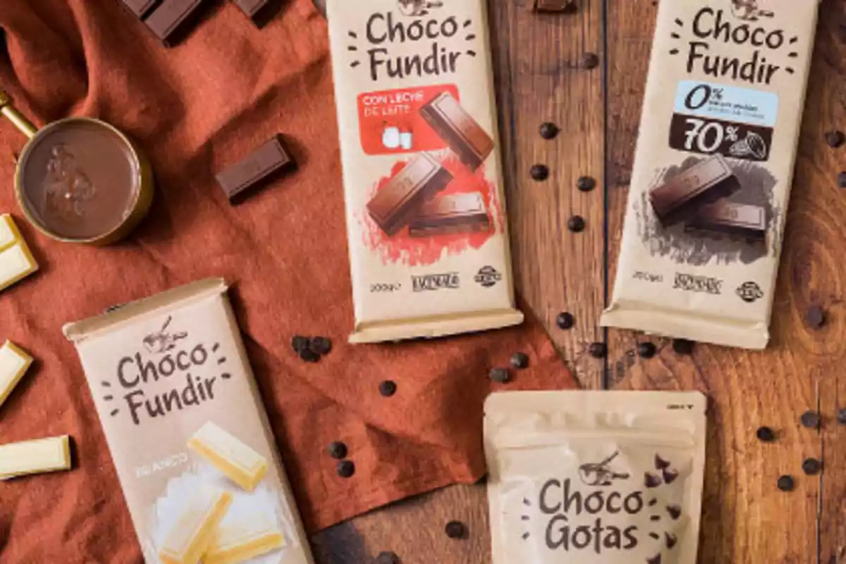 Vista superior de varias presentaciones de chocolate para fundir y chispas de chocolate sobre una mesa de madera y tela marrón, acompañadas de trozos de chocolate y una taza con chocolate derretido