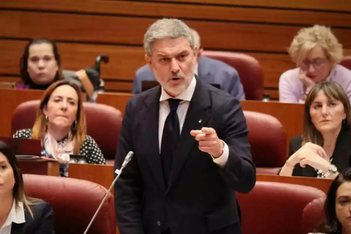 Hombre de cabello canoso y barba hablando enérgicamente y señalando con el dedo en una sala de reuniones con varias personas sentadas alrededor observando atentamente Hombre de cabello canoso y barba hablando enérgicamente y señalando con el dedo en una sala de reuniones con varias personas sentadas alrededor observando atentamente