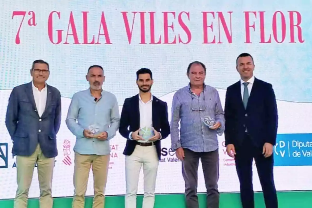 Cinco hombres posan en el escenario de un evento con el cartel de la 7ª Gala Viles en Flor, tres de ellos sostienen trofeos y todos visten ropa formal.