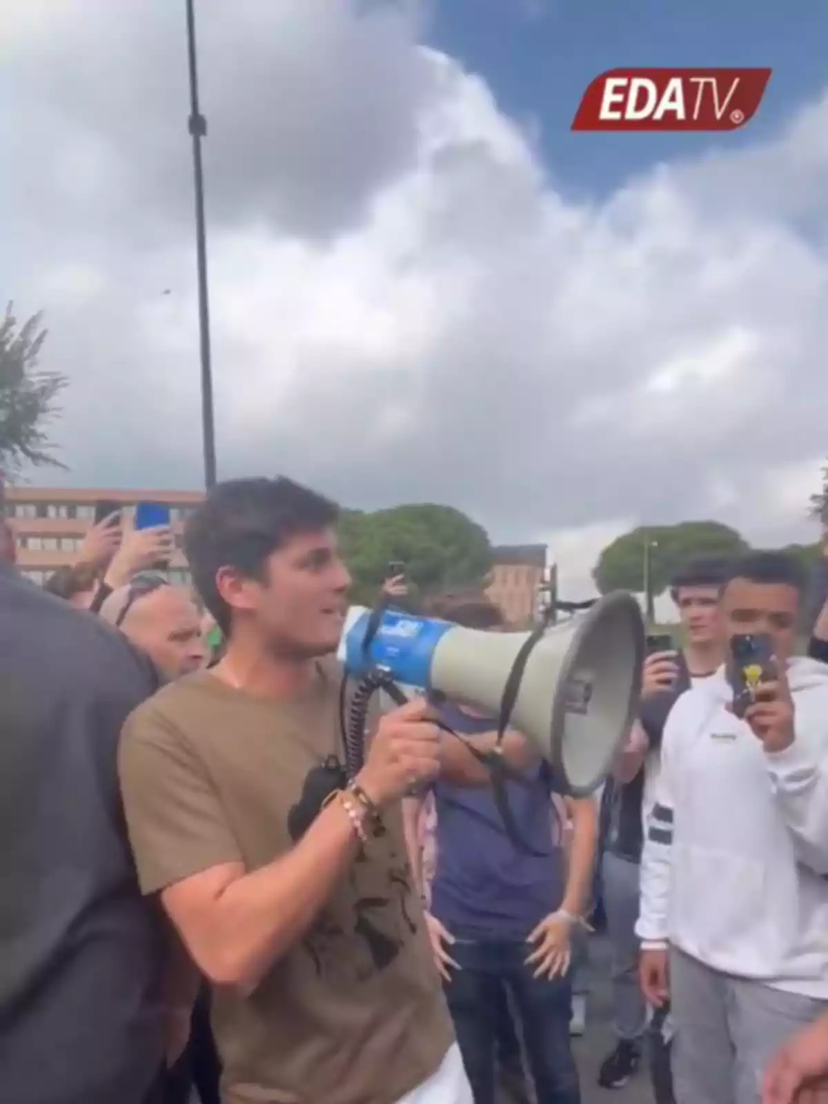 Un joven sostiene un megáfono mientras habla a un grupo de personas que lo rodean y lo graban con sus teléfonos móviles en un evento al aire libre bajo un cielo nublado.