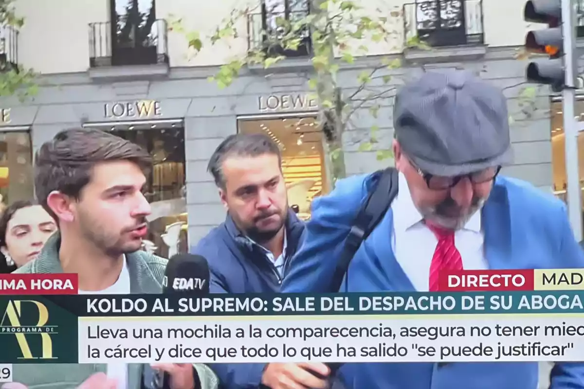 Un reportero entrevista a un hombre con gorra y gafas que lleva una mochila, mientras otra persona los acompaña en una calle comercial con tiendas de fondo y un rótulo informativo en la parte inferior de la imagen.