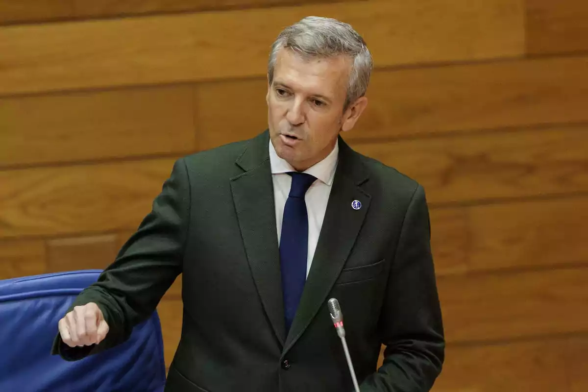 Hombre de cabello canoso con traje oscuro y corbata azul hablando frente a un micrófono en un entorno de madera