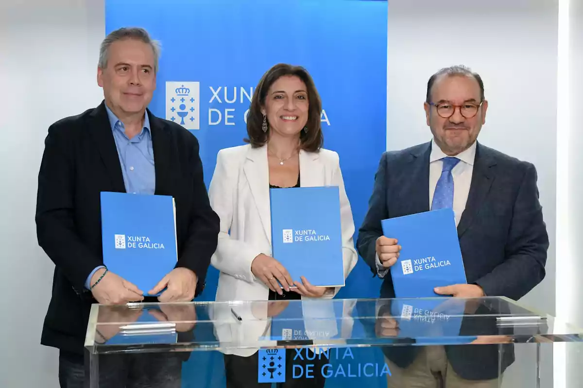 Tres personas posan sonrientes sosteniendo carpetas azules de la Xunta de Galicia frente a un atril y un fondo institucional azul