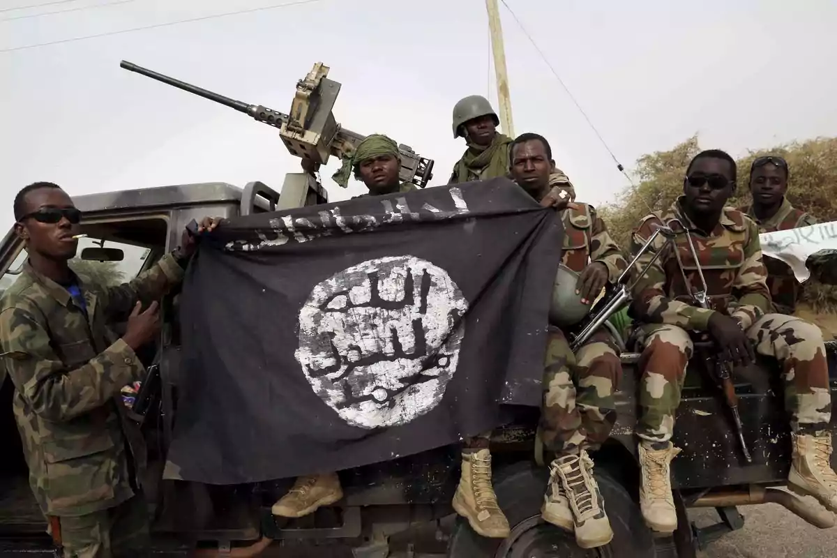 Soldados con uniforme de camuflaje sentados en un vehículo militar sosteniendo una bandera negra con inscripciones en árabe y un arma montada en el fondo