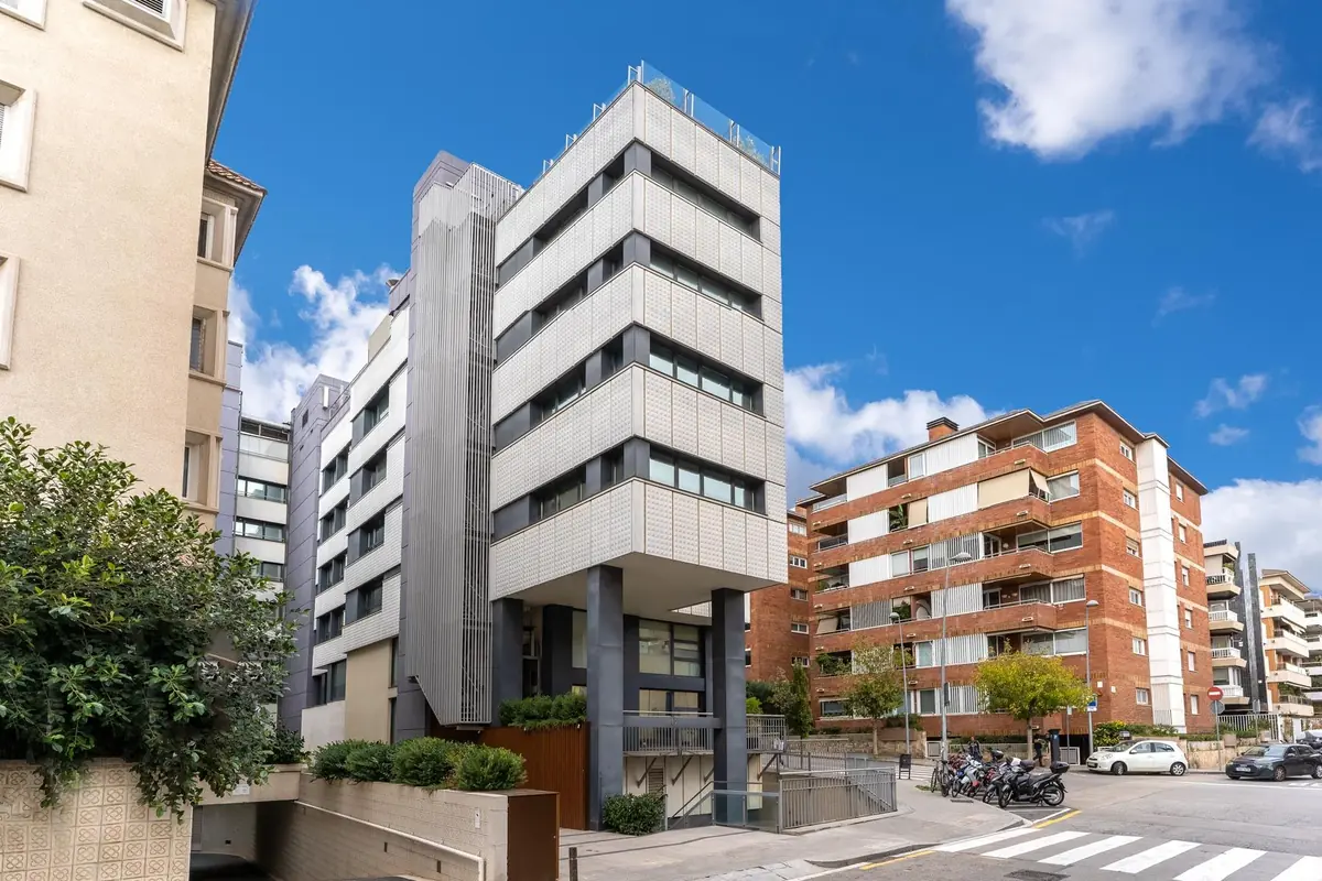 Edificio moderno de varios pisos con fachada de paneles claros y ventanales, ubicado en una esquina urbana junto a otros edificios residenciales y un cielo azul con algunas nubes
