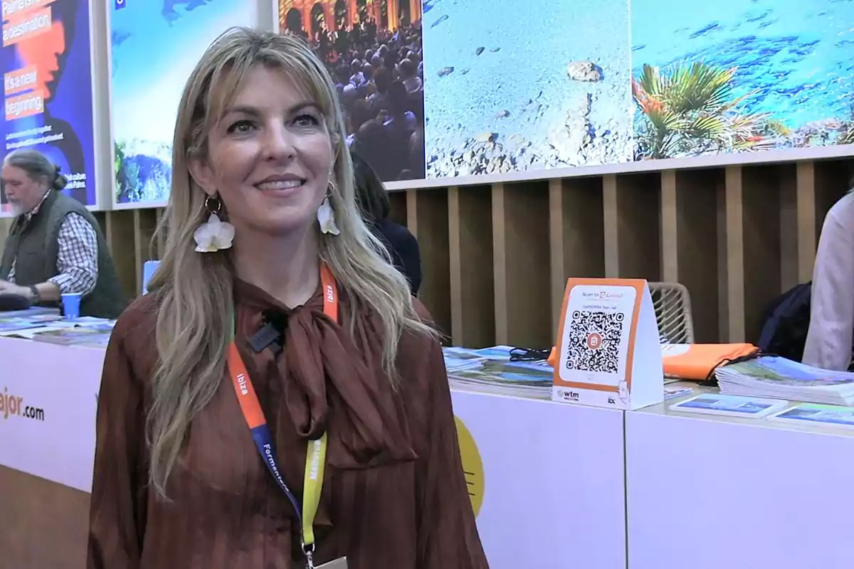 Mujer rubia con blusa marrón y aretes grandes sonríe en un stand de feria turística con folletos y un código QR sobre la mesa