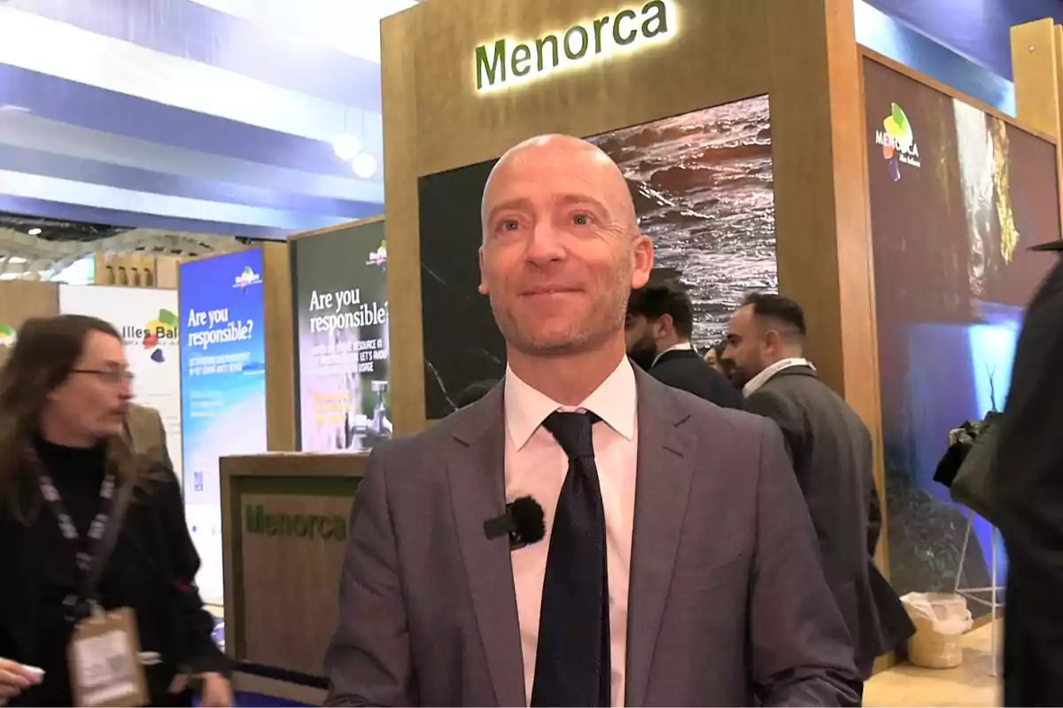 Hombre calvo con traje y corbata sonriendo en un stand de Menorca en una feria o evento, con varias personas y carteles de fondo.