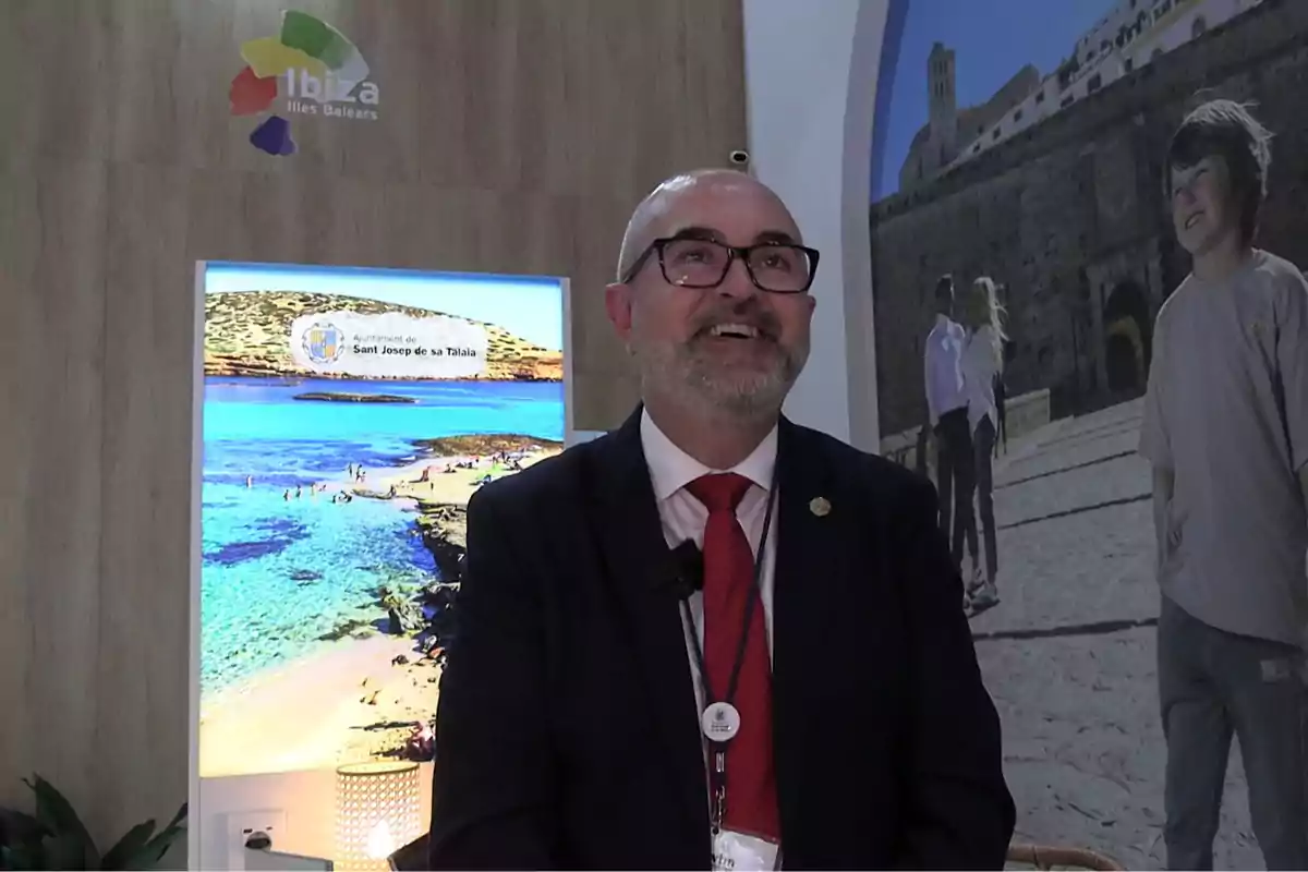 Hombre con gafas y barba canosa sonríe mientras viste traje oscuro y corbata roja en un stand promocional de Ibiza con imágenes turísticas de fondo