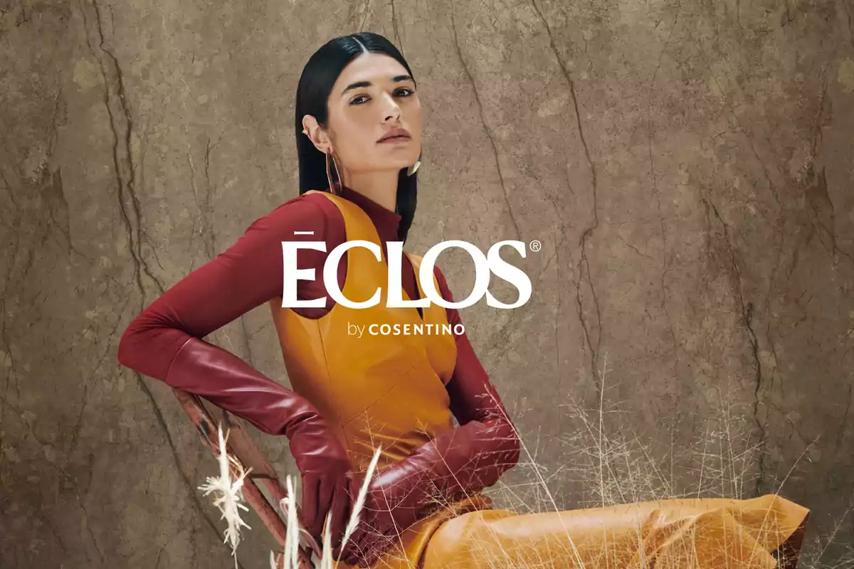 Mujer con cabello lacio y oscuro vestida con guantes rojos y un vestido naranja posando frente a un fondo de textura marmoleada con el texto ÉCLOS by Cosentino al centro