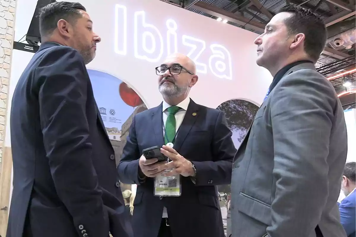Tres hombres de negocios conversan en un evento frente a un letrero luminoso que dice Ibiza
