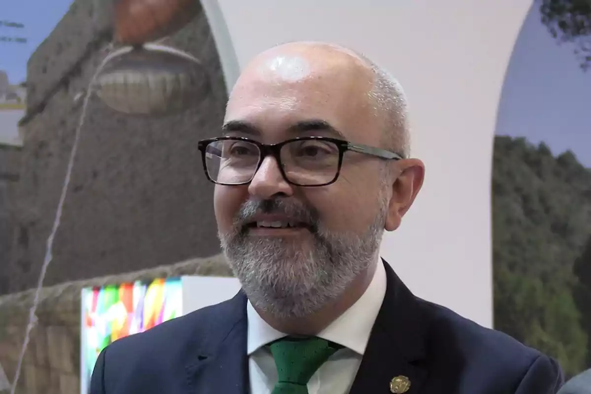 Hombre de mediana edad con barba y gafas, vestido con traje oscuro y corbata verde, sonríe en un entorno interior con fondo de imágenes de paisajes.