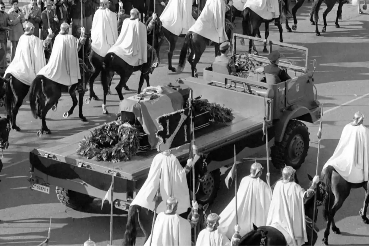 Desfile fúnebre con un ataúd cubierto por una bandera sobre un vehículo militar escoltado por jinetes vestidos con capas blancas y cascos