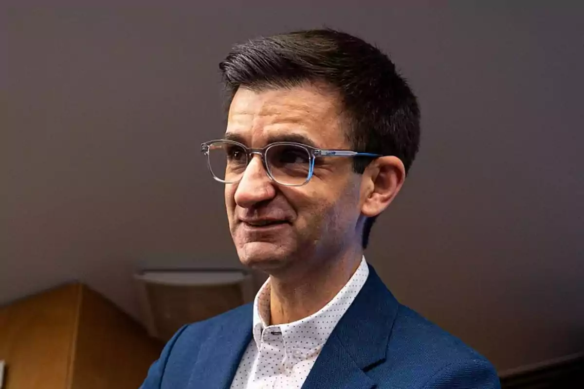 Hombre con gafas y cabello corto oscuro vistiendo saco azul y camisa blanca con puntos pequeños en un ambiente interior Hombre con gafas y cabello corto oscuro vistiendo saco azul y camisa blanca con puntos pequeños en un ambiente interior