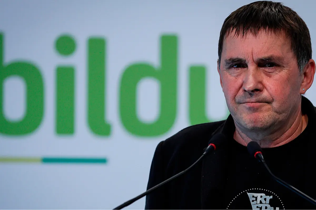 Hombre de cabello corto y oscuro hablando frente a dos micrófonos con el logo de Bildu desenfocado en el fondo