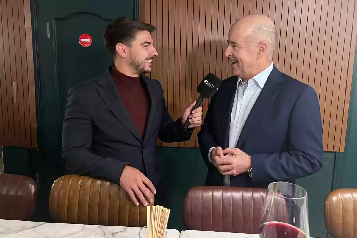 Un reportero entrevista a un hombre en un ambiente interior elegante mientras ambos sonríen y conversan, con una copa de vino y palillos sobre la mesa frente a ellos.