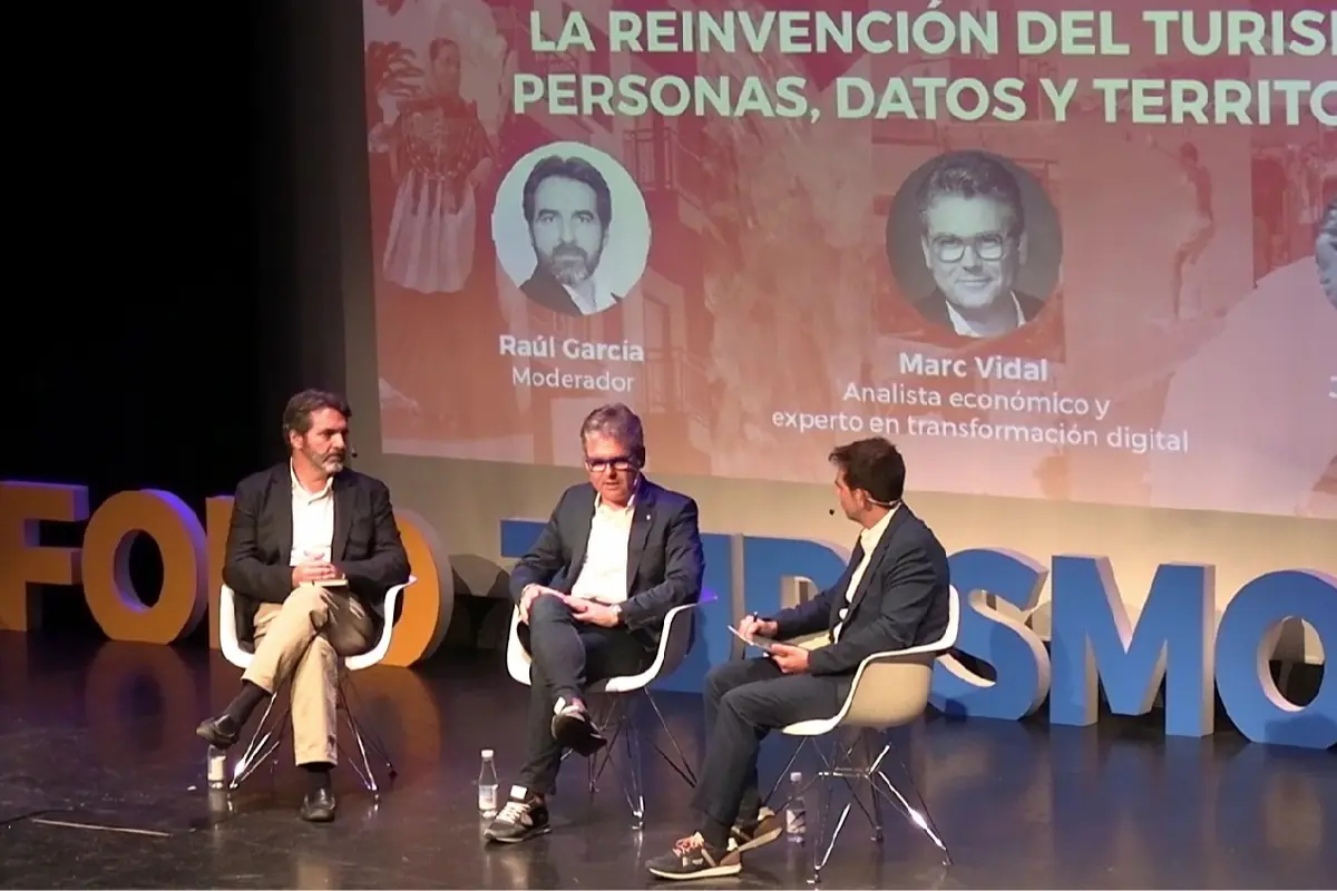 Tres hombres con ropa formal están sentados en sillas blancas sobre un escenario durante un foro sobre turismo, con letras grandes de colores en el fondo y una pantalla que muestra información del evento y los nombres de los ponentes.