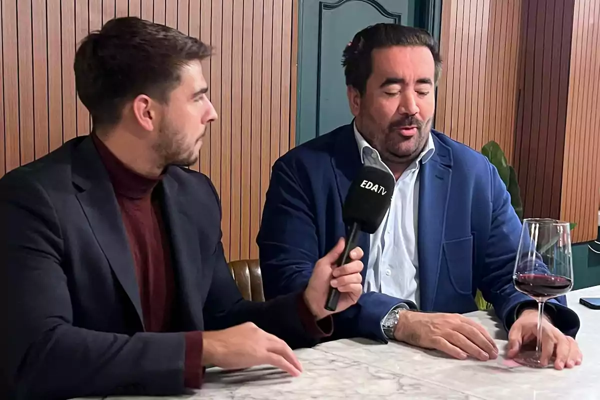 Dos hombres sentados en una mesa de mármol durante una entrevista, uno sostiene un micrófono con el logo de EDATV y el otro tiene una copa de vino tinto frente a él