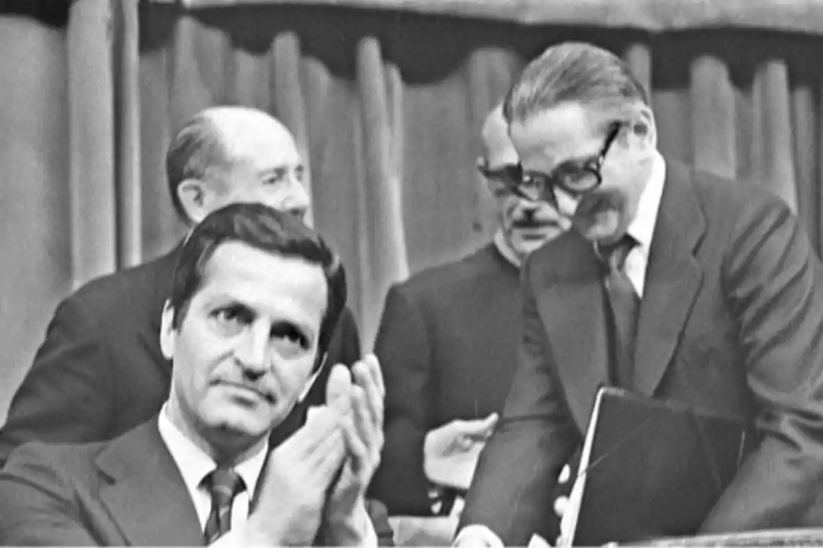 Cuatro hombres vestidos de traje en un ambiente formal, uno de ellos aplaudiendo y otro sonriendo mientras sostiene una carpeta