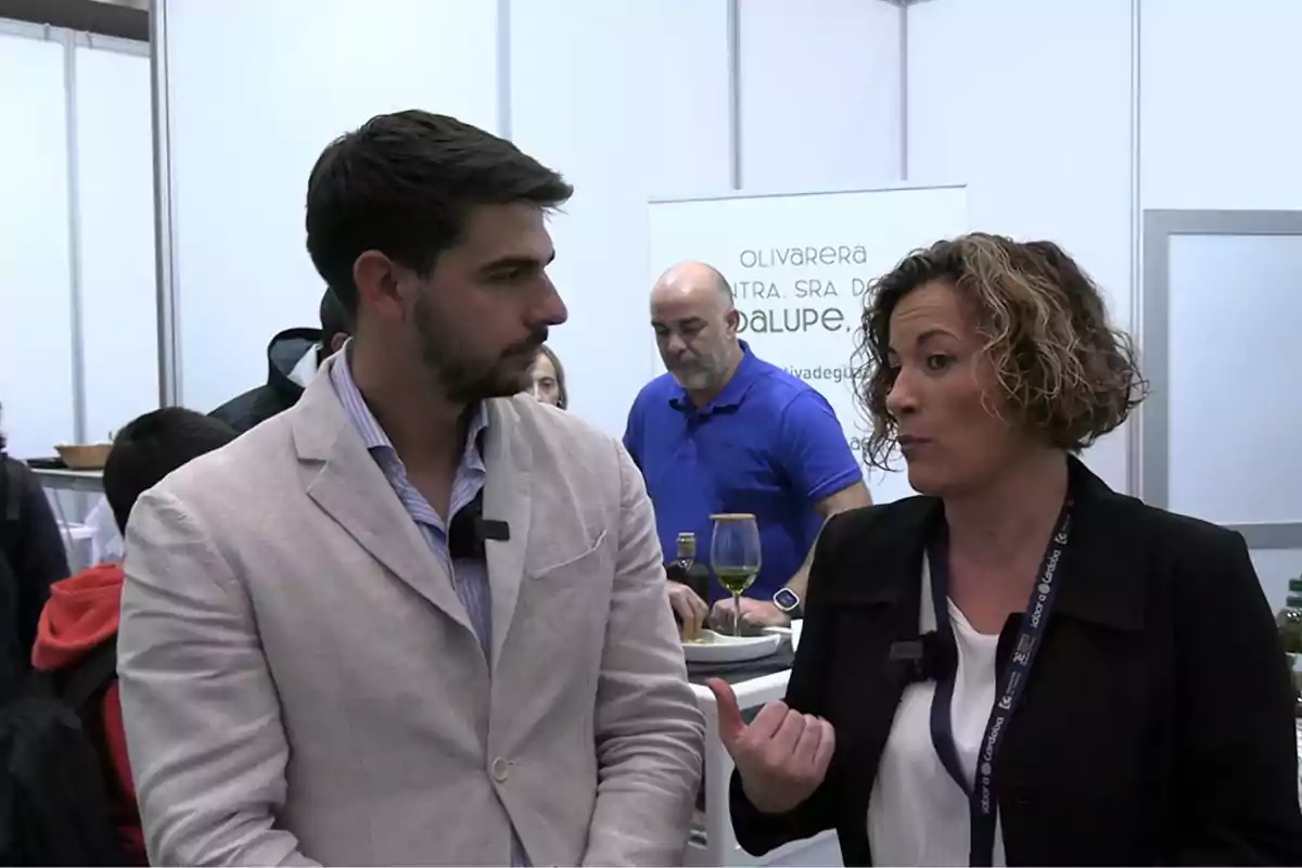 Dos personas conversan en un evento mientras al fondo se observa a un hombre sirviendo vino y un cartel promocional de una olivarera