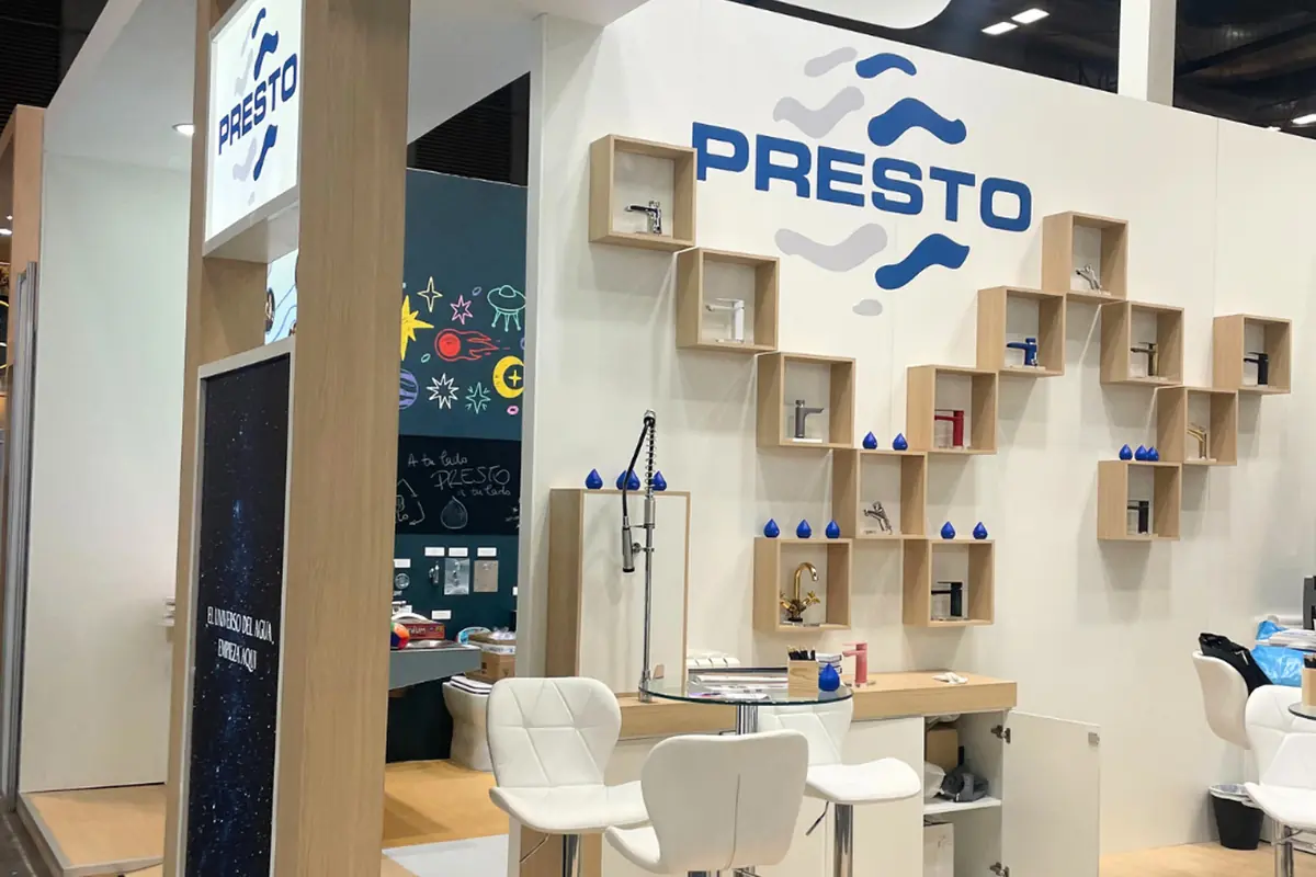 Stand de exhibición de la marca Presto con grifería expuesta en repisas de madera y sillas blancas frente a una mesa de cristal