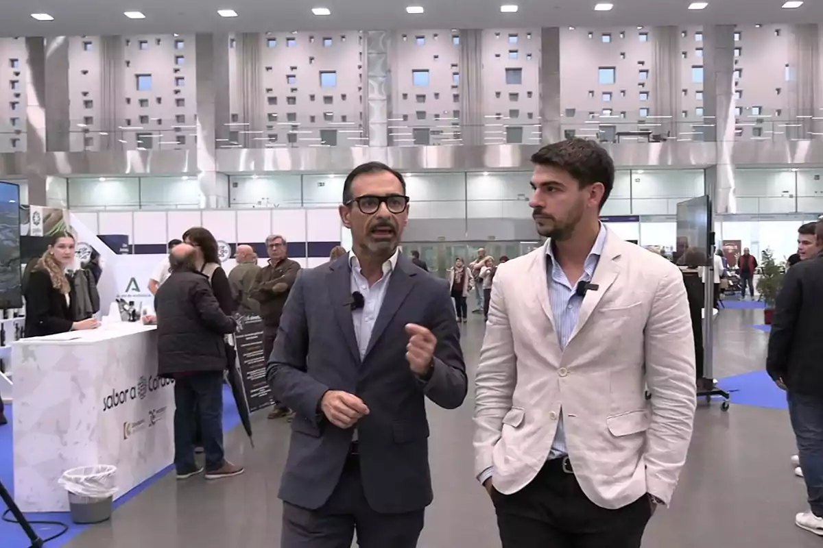 Dos hombres con micrófonos de solapa conversan en un evento o feria en un recinto moderno y luminoso con varias personas y stands al fondo
