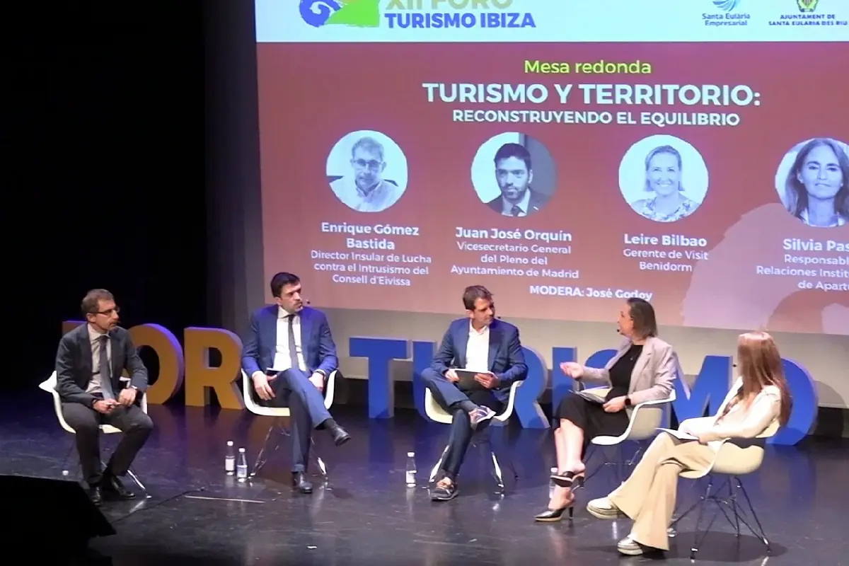 Cinco personas sentadas en sillas blancas participan en una mesa redonda sobre turismo y territorio en un escenario, con un fondo que muestra los nombres y cargos de los ponentes.