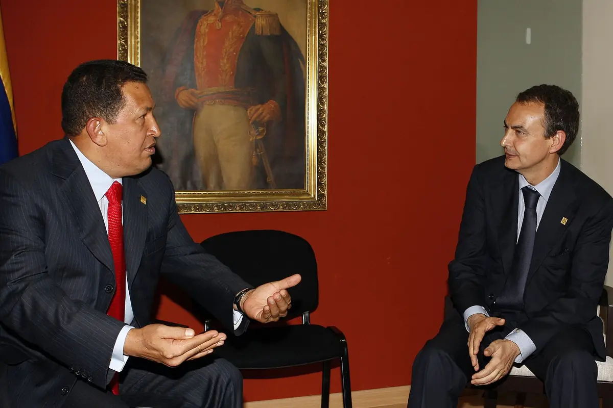 Dos hombres con traje conversan sentados en una sala con una pared roja y un retrato de un militar al fondo Dos hombres con traje conversan sentados en una sala con una pared roja y un retrato de un militar al fondo