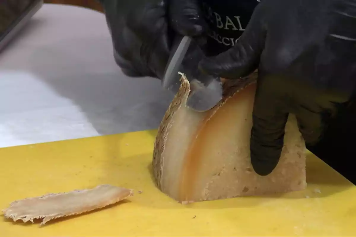 Manos con guantes negros cortando una cuña de queso curado sobre una tabla amarilla