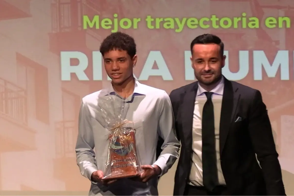 Dos personas posan en un escenario durante una ceremonia de premiación, una de ellas sostiene un trofeo envuelto en celofán y al fondo se puede leer parte de un texto que dice Mejor trayectoria