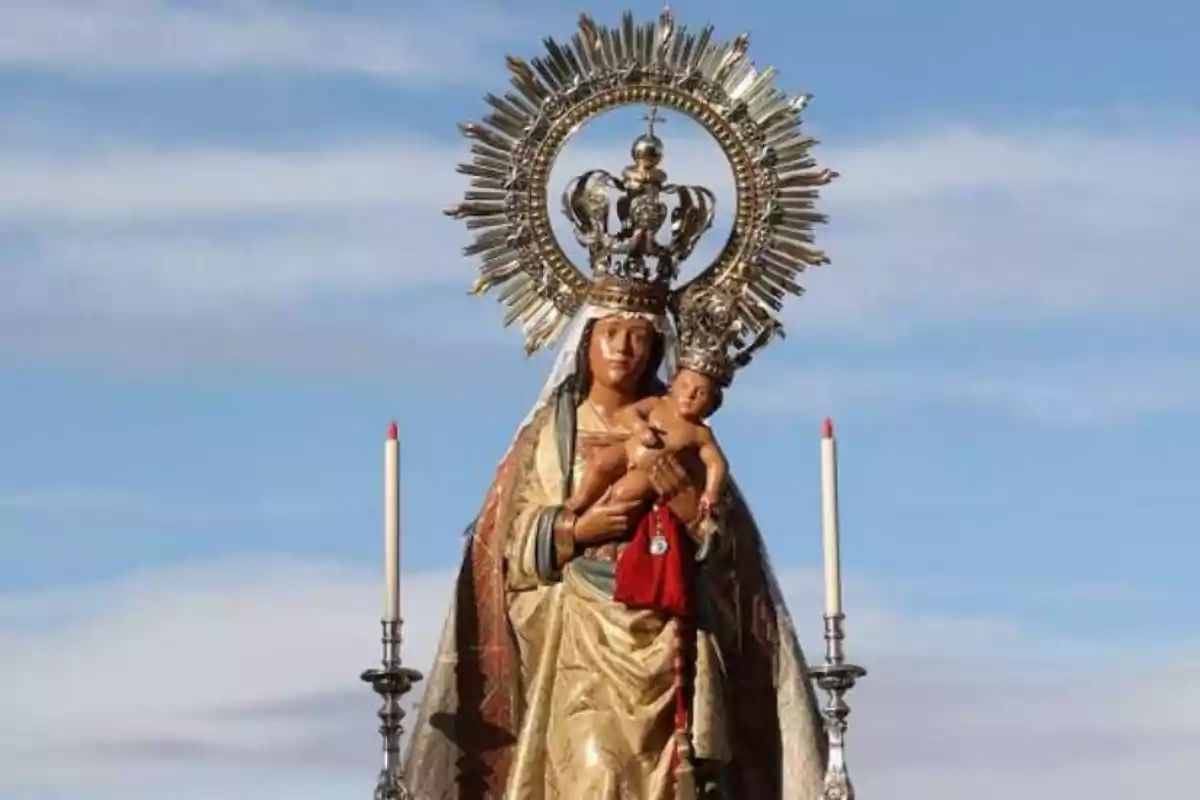 Imagen de una estatua religiosa de la Virgen con el Niño Jesús en brazos ambos con coronas y un resplandor metálico detrás de la Virgen acompañados por dos velas a los lados bajo un cielo azul.