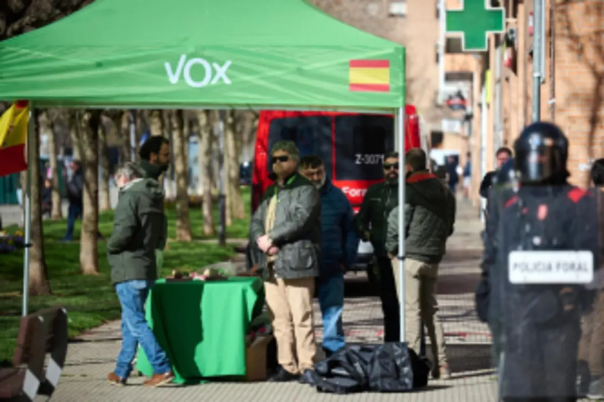 Varias personas se encuentran reunidas bajo una carpa verde de VOX en la calle mientras un agente de la policía foral con escudo observa la escena