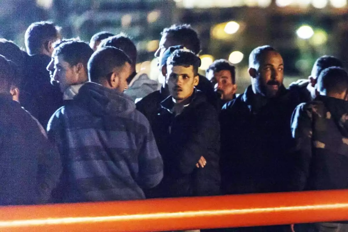 Un grupo de personas de pie en la noche con expresiones serias y luces desenfocadas al fondo