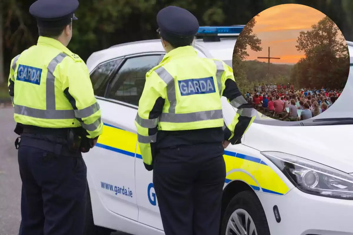 Dos agentes de policía irlandesa de espaldas junto a un coche patrulla con un recuadro que muestra a un grupo de personas reunidas al aire libre frente a una cruz al atardecer