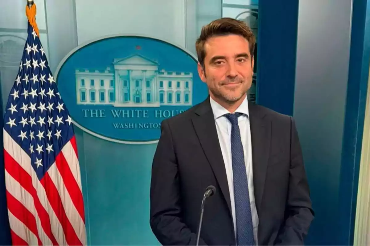 Hombre con traje y corbata posando frente a un podio en la sala de prensa de la Casa Blanca junto a una bandera de Estados Unidos