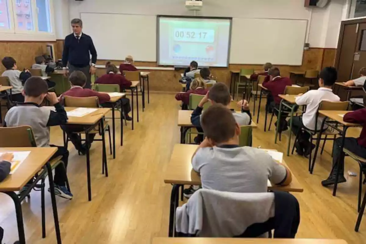 Niños sentados en pupitres individuales en un aula mientras un maestro supervisa y un cronómetro se proyecta en la pizarra.