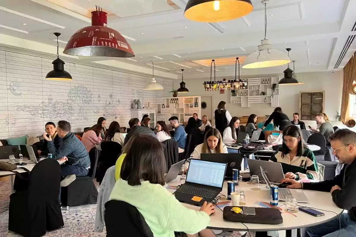 Varias personas trabajan en computadoras portátiles alrededor de mesas en un espacio moderno y bien iluminado, decorado con lámparas colgantes y una pared con dibujos.