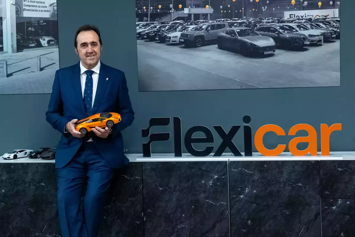 Hombre de traje azul sosteniendo un auto de juguete naranja junto al logo de Flexicar en una oficina con fotos de autos en la pared