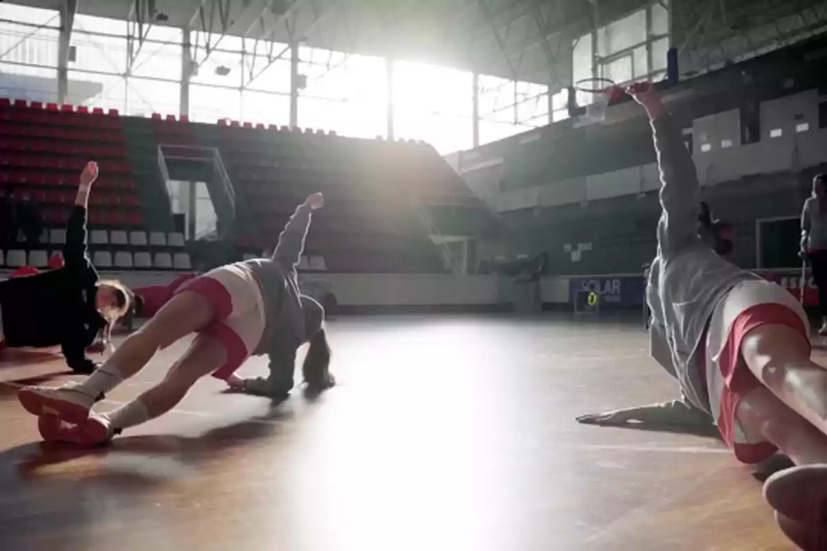 Jugadoras de baloncesto realizando ejercicios de estiramiento en una cancha techada iluminada por la luz del sol