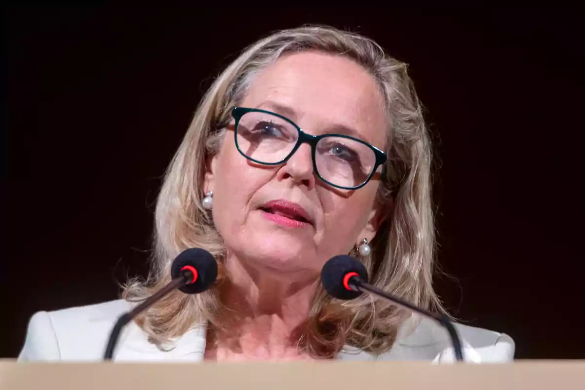Mujer rubia con gafas y chaqueta clara hablando frente a dos micrófonos