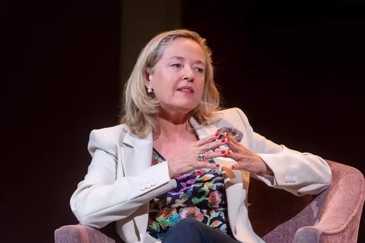 Mujer sentada en un sillón con chaqueta clara y blusa colorida gesticulando con las manos mientras participa en una conversación