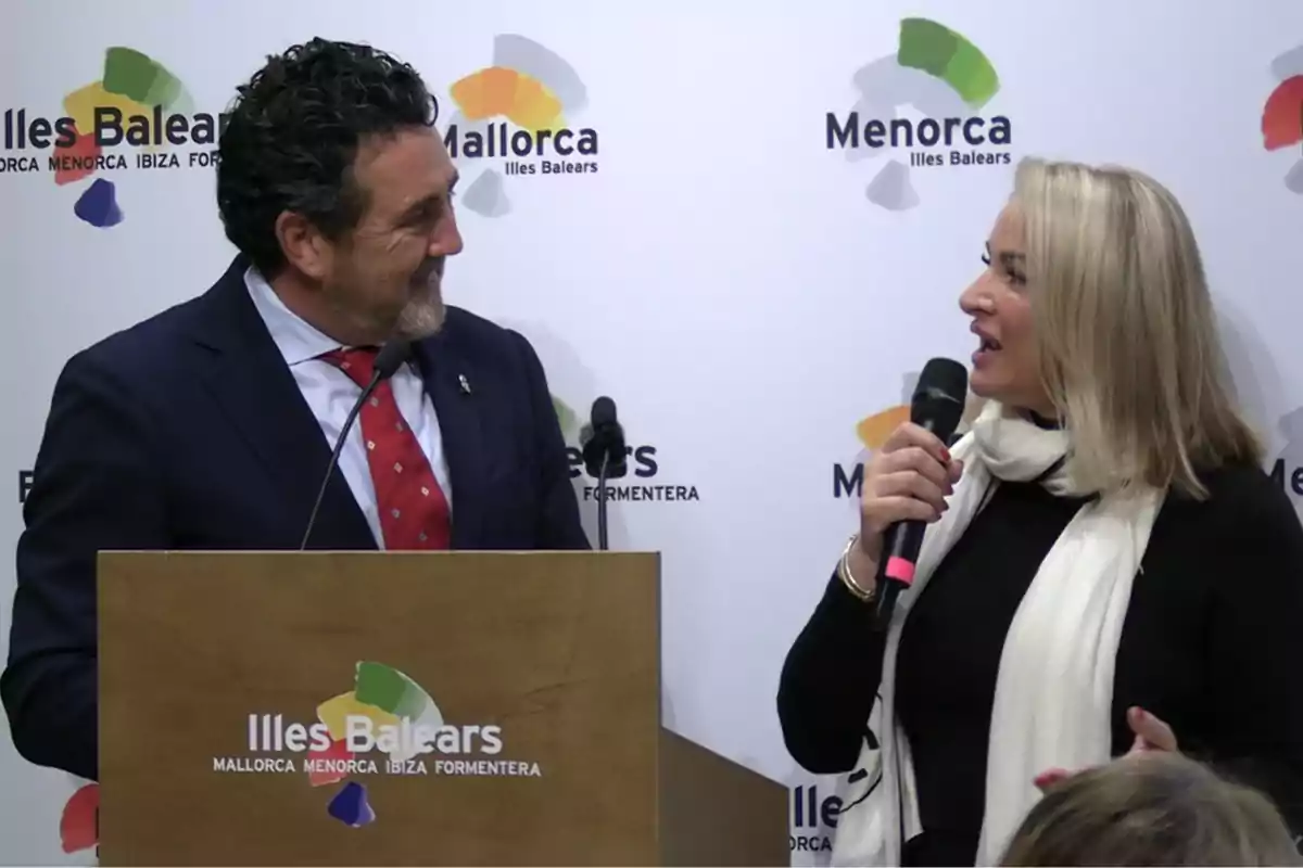 Un hombre con traje y una mujer con bufanda blanca conversan sonrientes en un evento frente a un atril con el logo de Illes Balears