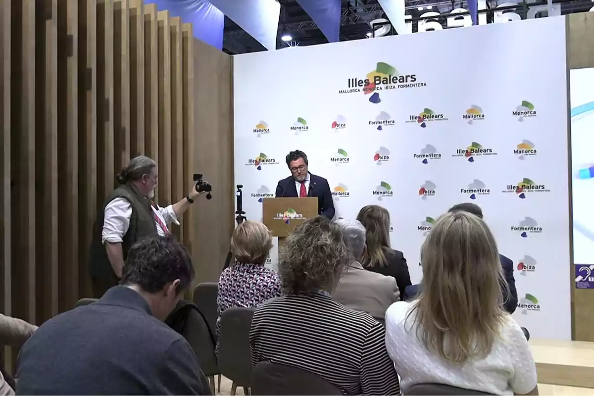 Un hombre de traje da un discurso en un podio frente a una audiencia mientras un fotógrafo lo graba en un evento promocional de las Islas Baleares