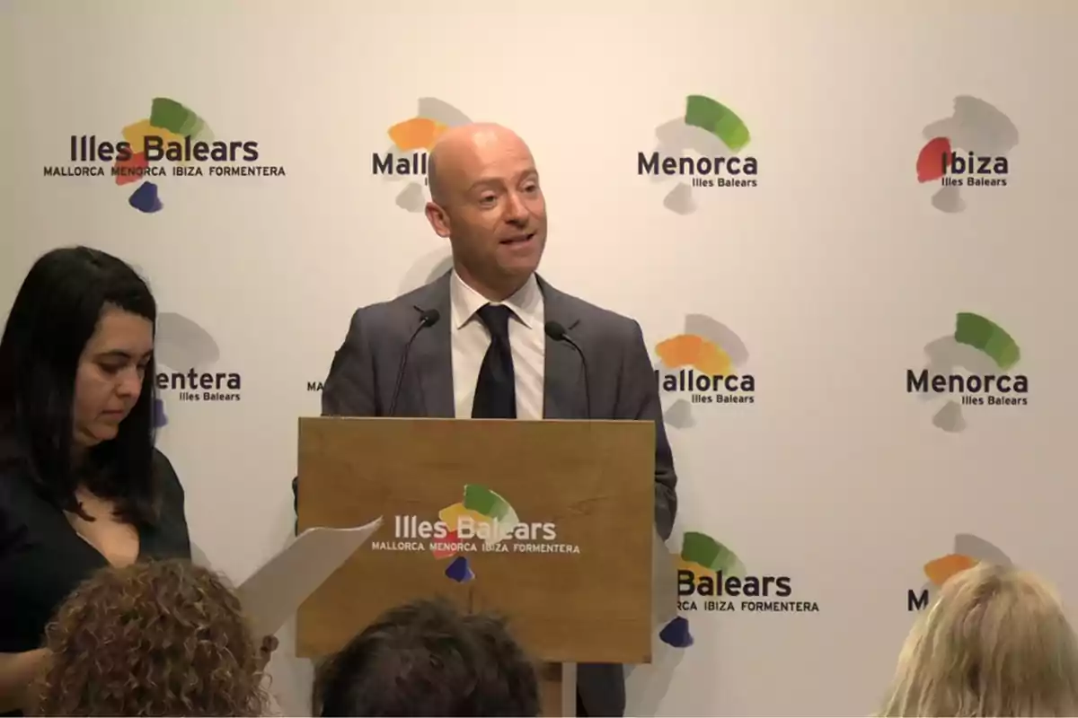 Un hombre con traje y corbata habla en un atril durante una conferencia de prensa de las Islas Baleares mientras una mujer sostiene unos papeles a su lado y varias personas lo escuchan.